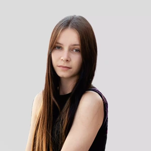 Portrait photo of a young woman with long straight hair, wearing a black sleeveless top. She is looking at the camera with a serious expression and has a nose ring. The background is neutral light grey.