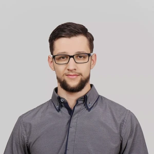 Portrait photo of a man wearing glasses, with short dark hair and a beard. He is dressed in a grey checkered shirt with a dark blue trim. The background is light grey.