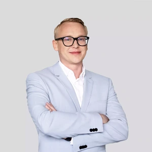Portrait photo of a man with short blond hair, wearing glasses, a light grey suit, and a white shirt. He is standing with arms crossed, smiling and looking at the camera against a neutral background.