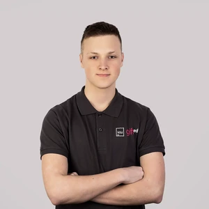 Young man with short hair, wearing a dark shirt with the “KTU GIFTed” logo, standing with arms crossed against a gray background.