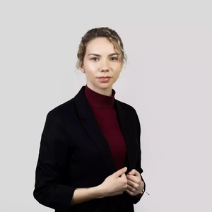 Portrait photo of a woman with light wavy hair tied back in a bun. She is wearing a dark blazer and a burgundy turtleneck. The background is light grey with a subtle pinkish tone on the right side.