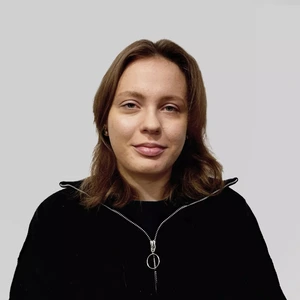 Portrait photo of a young woman with brown hair, wearing a black zip-up sweater with a circular zipper pull. She is looking at the camera against a neutral gray background.