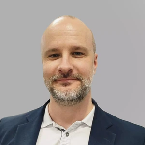 Middle-aged man with a beard, wearing a light shirt and a dark blazer, smiling in front of a gray background.