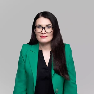 Portrait photo of a woman with long dark hair, wearing a bright green blazer and a black blouse. She is looking straight at the camera through glasses with a gentle smile. The background is neutral light grey.