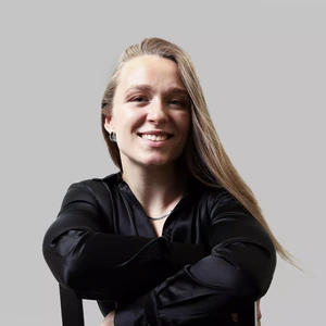 Portrait photo of a young woman with long blond hair, wearing a black jacket with long sleeves. She is looking at the camera with a broad smile, arms crossed, against a neutral grey background.