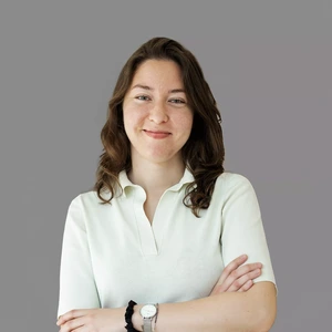 Portrait photo of a young woman with curly dark hair, wearing a light-colored blouse with short sleeves. She is looking at the camera with a gentle smile, arms crossed, against a neutral grey background.