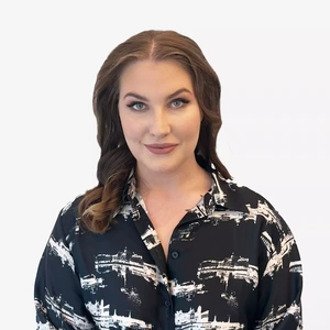 A young woman with wavy light brown hair, wearing a black shirt with a white abstract pattern, posing against a light background.