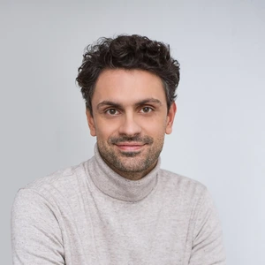 Portrait photo of a man with curly dark hair, wearing a grey sweater with a high collar. He is looking at the camera with a gentle smile, against a neutral grey background.