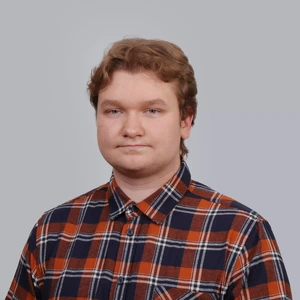 Young man wearing a plaid shirt in orange and dark blue tones, standing in front of a gray background and looking directly at the camera.