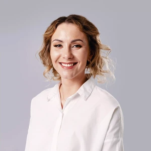 Portrait photo of a woman with light, curly hair styled loosely. She is wearing a white shirt and smiling. The background is light grey and uniform.