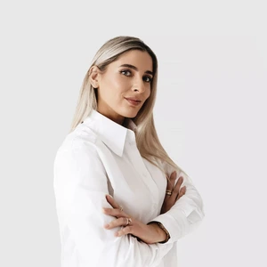 Portrait photo of a woman with long blonde hair, wearing a white shirt. She is standing sideways with arms crossed, looking at the camera confidently. The background is neutral light grey.