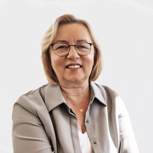 Mature woman with short blonde hair, wearing glasses, a light grey blazer, and a white shirt, smiling in front of a light background.