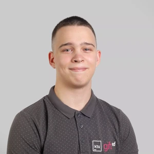 Portrait photo of a young man with short hair, wearing a dark grey polo shirt with the "ktu gifted" logo. He is looking at the camera with a slight smile, against a neutral grey background.