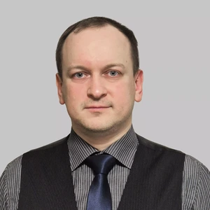 Portrait photo of a man with short dark hair, wearing striped shirt, dark vest, and a shiny blue tie. He is looking seriously at the camera. The background is neutral grey.