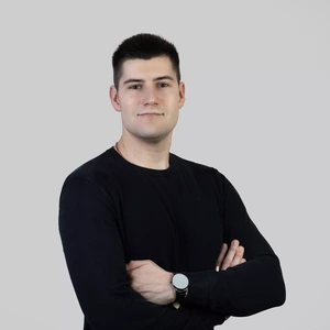 Portrait photo of a man with short dark hair, wearing a black sweater. His arms are crossed, and he's wearing a watch on his left wrist. The background is light grey and neutral.