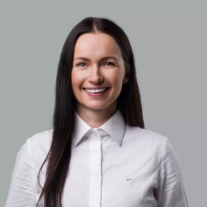 Portrait photo of a woman with long dark hair, wearing a white shirt. She is smiling broadly and looking directly at the camera. The background is a neutral grey-green.