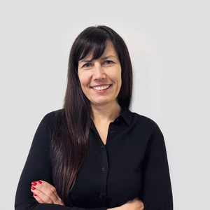 Portrait photo of a woman with long dark hair, wearing a black long-sleeve shirt. She is smiling with her arms crossed over her chest, against a neutral gray background.