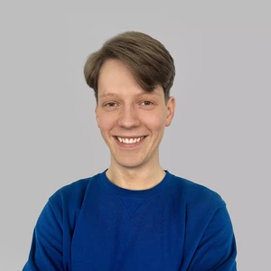 Portrait photo of a young man with light hair, wearing a blue sweater, smiling broadly and looking at the camera against a light background.