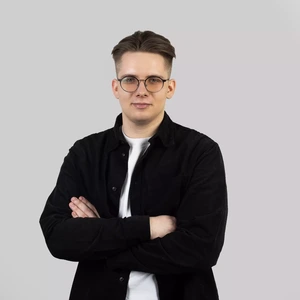 Portrait photo of a young man with short light brown hair, wearing round glasses. He is dressed in a white T-shirt and a black overshirt. His arms are crossed. The background is light grey.
