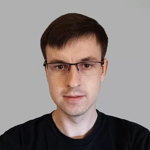 Portrait photo of a young man with dark hair and glasses, wearing a black t-shirt-style top, looking directly at the camera against a light background.