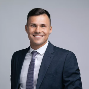 A young man with short dark hair, wearing a dark blue suit, white shirt, and patterned tie, is smiling broadly and looking at the camera against a light background.