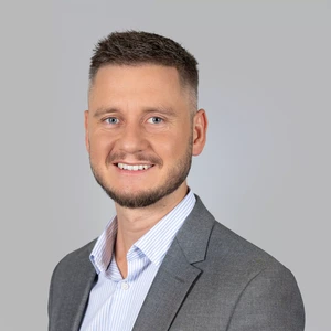 Portrait photo of a man with short light hair and a short beard, wearing a gray blazer and striped shirt. He is smiling and looking at the camera against a gray background.