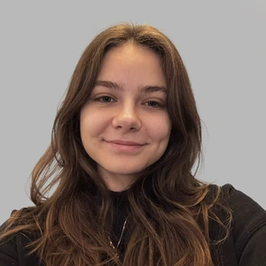 Portrait photo of a young woman with long brown hair, wearing black clothing, gently smiling while looking at the camera against a light background.