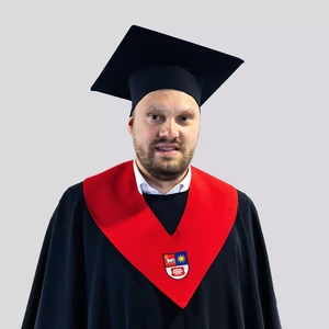Portrait photo of a man wearing academic regalia: a dark gown with a bright red collar featuring a coat of arms, and a dark academic cap. He is looking at the camera with a serious expression. The background is neutral light grey.