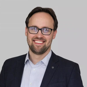 Portrait photo of a young man with a beard and glasses, wearing a dark blue suit and light checkered shirt. He is smiling and looking at the camera against a neutral gray background.