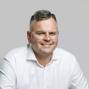 Portrait photo of a man with short greying hair, wearing a white shirt and metal-rimmed glasses. He is smiling while looking at the camera, against a neutral light grey background.