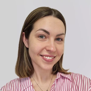 Portrait photo of a woman with short light brown hair, wearing a pink striped shirt. She is smiling and looking directly at the camera. The background is light and neutral.