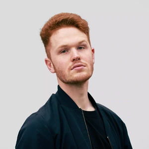 Portrait photo of a young man with red hair and a beard, wearing a black jacket and t-shirt, looking at the camera with a serious expression against a light background.