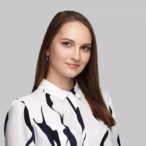 A young woman with straight brown hair, wearing a white shirt with black abstract patterns, looks at the camera with a soft smile against a light background.
