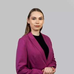 Portrait photo of a young woman with straight light hair, wearing a bright fuchsia blazer and a black top. She is looking directly at the camera with a confident, composed expression, hands positioned in front. The background is neutral light grey.