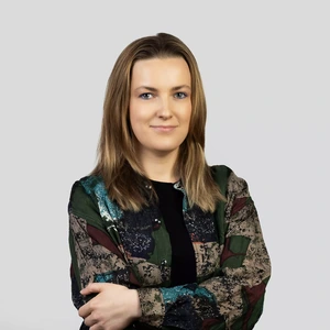 Portrait photo of a young woman with straight light brown hair, wearing a patterned blouse in green, black, and brown tones. She is standing with arms crossed and looking at the camera with a slight smile. The background is neutral light grey.