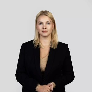 Portrait photo of a young woman with straight blonde hair, wearing a black blazer and a beige top. She is standing straight and looking at the camera with a serious yet calm expression. The background is light grey and neutral.