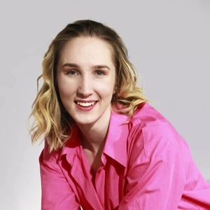 Portrait photo of a woman with wavy blonde hair, wearing a bright pink blouse. She is smiling and looking directly at the camera. The background is light grey.