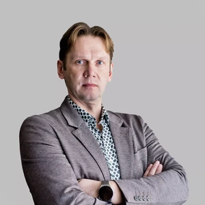 Middle-aged man with light hair, wearing a brown blazer and patterned shirt, standing with arms crossed and a serious expression, light grey background.