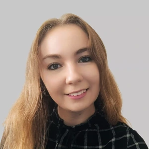 Young woman with light, medium-length hair, wearing a black checkered shirt, smiling and looking at the camera, grey background.