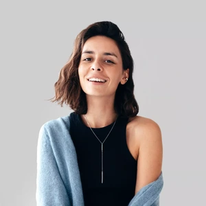 Young woman with short dark wavy hair, wearing a black sleeveless top and a light blue sweater draped over her shoulders, smiling at the camera, grey background.
