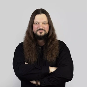 Portrait photo of a man with long dark hair and a beard, wearing a black shirt. He is standing with arms crossed and looking at the camera with a slight smile. The background is neutral light grey.
