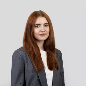 Portrait photo of a woman with long straight copper-red hair, wearing a grey blazer with decorative elements and a white top. She is looking at the camera with a subtle smile. The background is light grey.