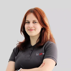 Portrait photo of a young woman with wavy copper-colored hair, wearing a dark grey polo shirt with the "ktu gifted" logo. She is smiling, arms crossed. The background is light grey.