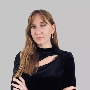 Serious-looking woman with long light brown hair, wearing a black velvet top with a cutout design and pearl-like earrings, grey background.