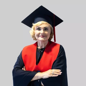 Portrait photo of a woman with light curly hair, wearing academic regalia with a bright red collar accent and a dark academic cap with a red tassel. She is standing with arms crossed and smiling at the camera. The background is neutral light grey.