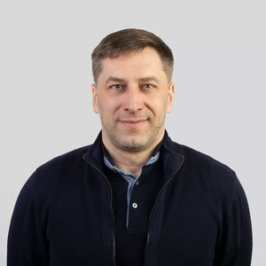 Portrait photo of a man with short light hair, wearing a dark blue zip-up sweater and light shirt. He is looking at the camera with a gentle smile, against a neutral grey background.