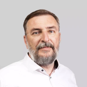 Portrait photo of an older man with a full beard, wearing white shirt. He is looking at the camera with a serious expression, against a neutral grey background.