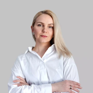 A woman with blonde hair, wearing a white shirt, stands with arms crossed and looks at the camera with a confident, serious expression against a light background.