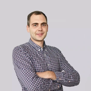 Portrait photo of a young man with short hair, wearing dark checkered long-sleeved shirt. He is standing with his arms crossed and looking at the camera against a neutral gray background.