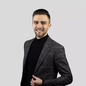 Portrait photo of a young man with light hair and a beard, wearing a dark checkered blazer and a black turtleneck. He is smiling and looking at the camera against a neutral gray background, holding one side of his blazer with his hand.